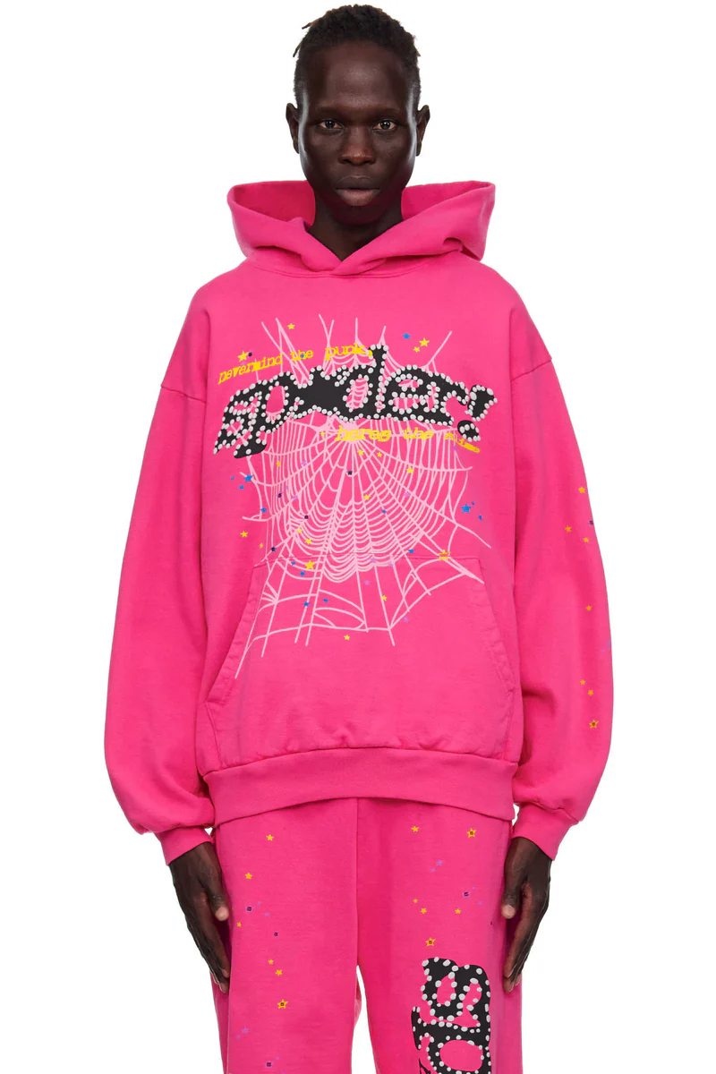 Model in a hot magenta Sp5der hoodie and matching sweatpants, with a black studded wordmark over a pale pink spider web, yellow typewriter text and small yellow and blue stars
