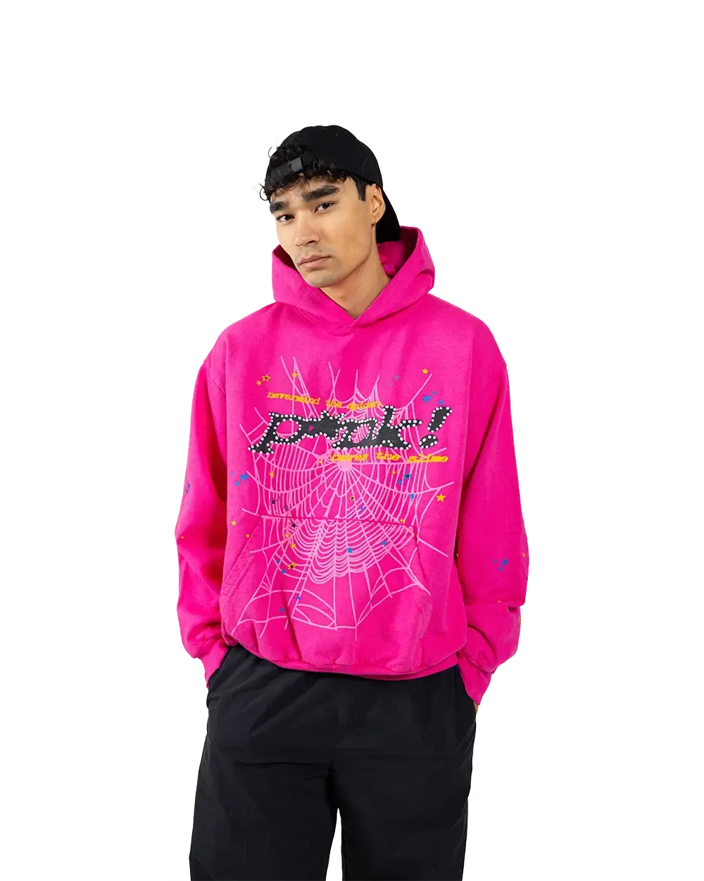 Man wearing a hot magenta Sp5der P*NK V2 hoodie with a black studded P*NK logo, yellow typewriter text and a pale pink web across the chest, paired with black trousers and a backwards black cap