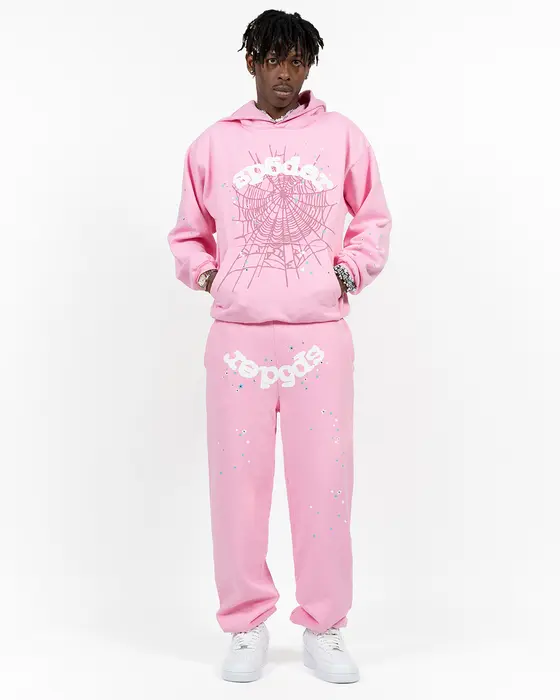 Man in a soft pink Sp5der OG Web hoodie and matching pink sweatpants, both printed with white puff sp5der lettering, the hoodie hanging loose past the waist and the sweatpants stacked over white sneakers