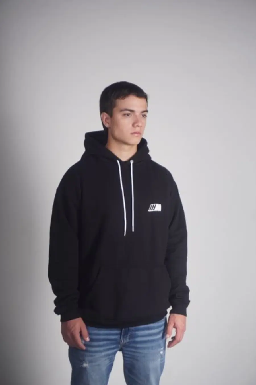 Portrait of a young man wearing a black hoodie and jeans against a gray backdrop.