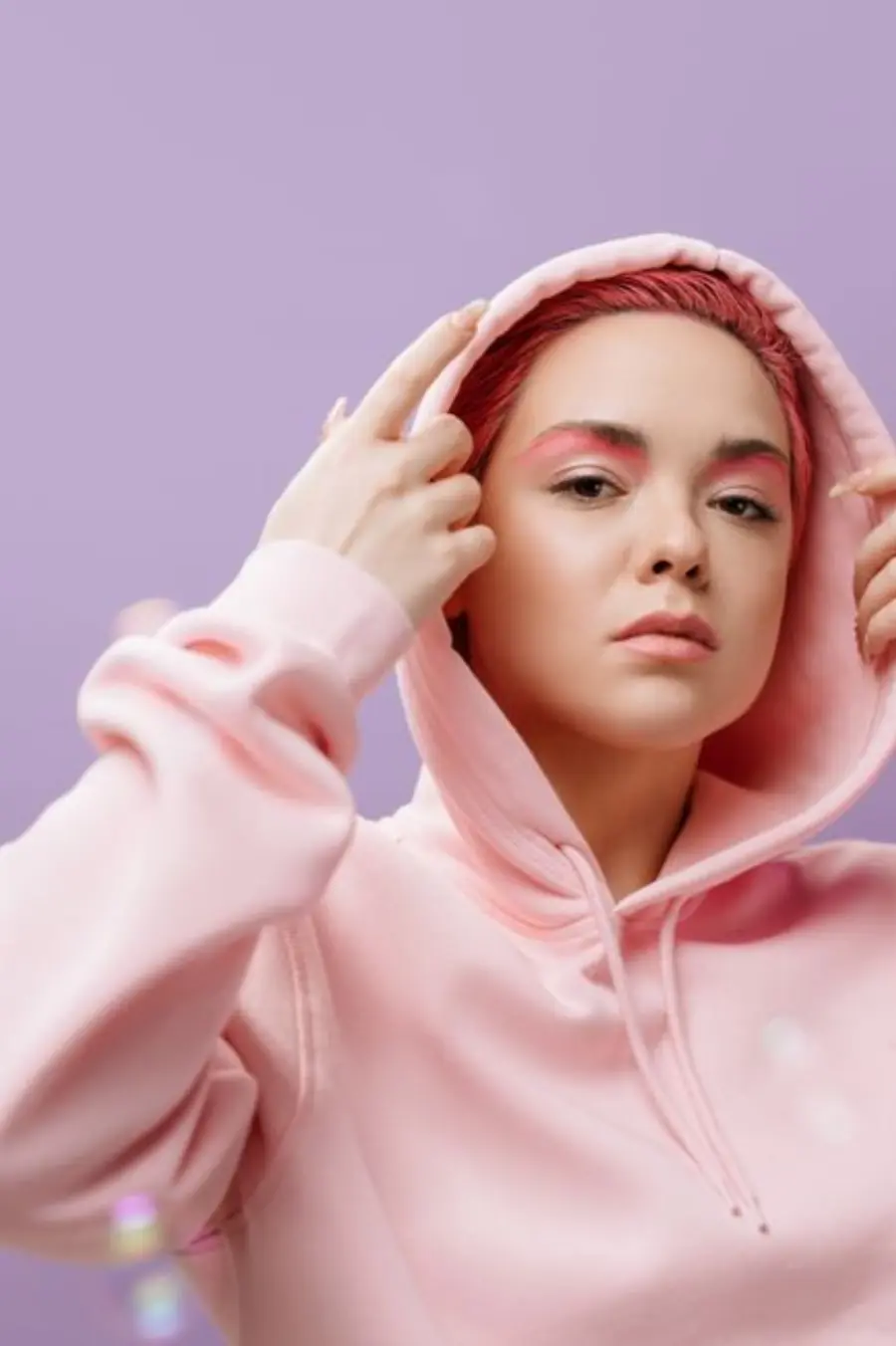 Modern portrait capturing a woman with pink hair and hoodie against a soft lilac background.